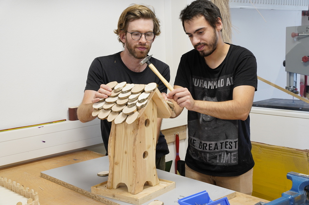 2 men working on a wooden birdhouse              