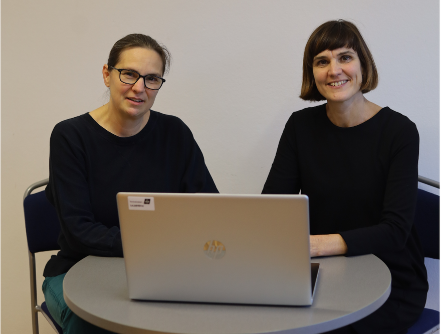 Das Foto zeigt zwei Frauen, die hinter einem Laptop sitzen.