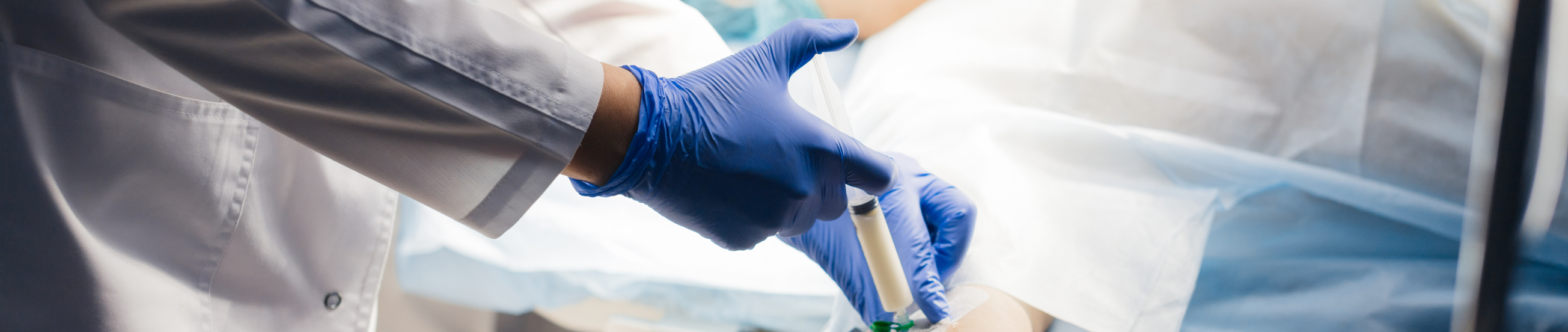 Male therapist puts a syringe with liquid medicine into an intravenous catheter, close up. Patient lyes in emergency intensive intensive care unit.