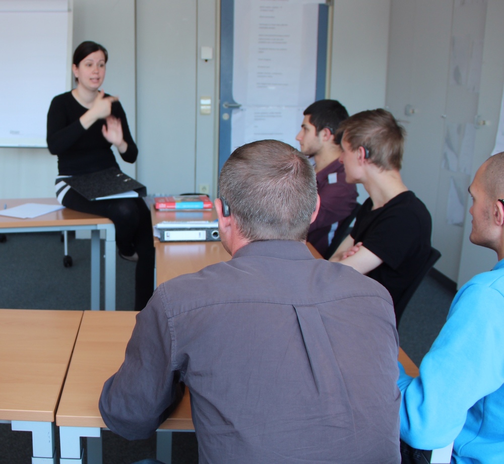 Das Bild zeigt Personen, welche einer Frau zusehen, die Gebärdensprache spricht.