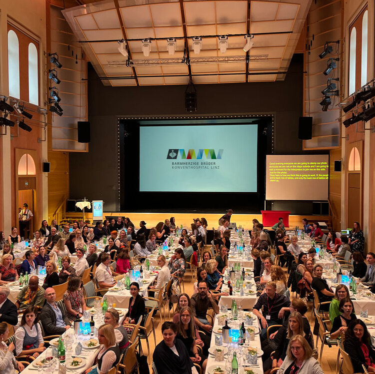 Das Foto zeigt einen großen Saal mit sehr vielen Menschen, die bei Tischen sitzen. Im Hintergrund ist ein Beamer mit dem Logo der Barmherzigen Brüder zu sehen.