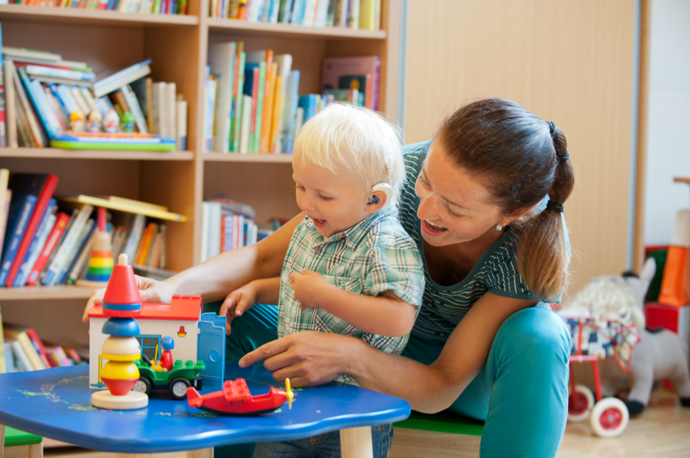 Das Foto zeigt eine Frau, die mit einem hörbeeinträchtigten kleinen blonden Jungen spielt.