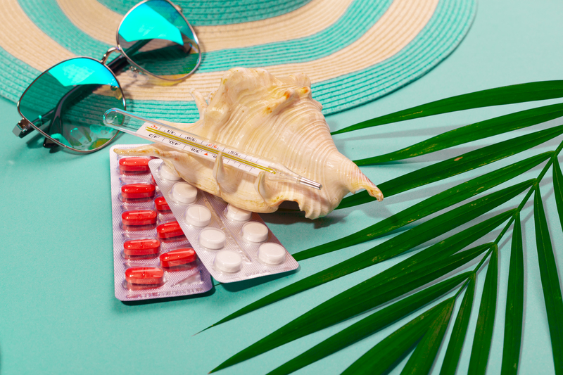 Top view on white and red fever pills and a thermometer in seashell, striped hat, sunglasses, blue flip-flops, palm leaf, on mint green background. Concept of preparing for sickness on vacation at sea. Advertising space Top view on white and red fever pills and a thermometer in seashell, striped hat, sunglasses, blue flip-flops, palm leaf, on mint green background. Concept of preparing for sickness on vacation at sea. Advertising space.