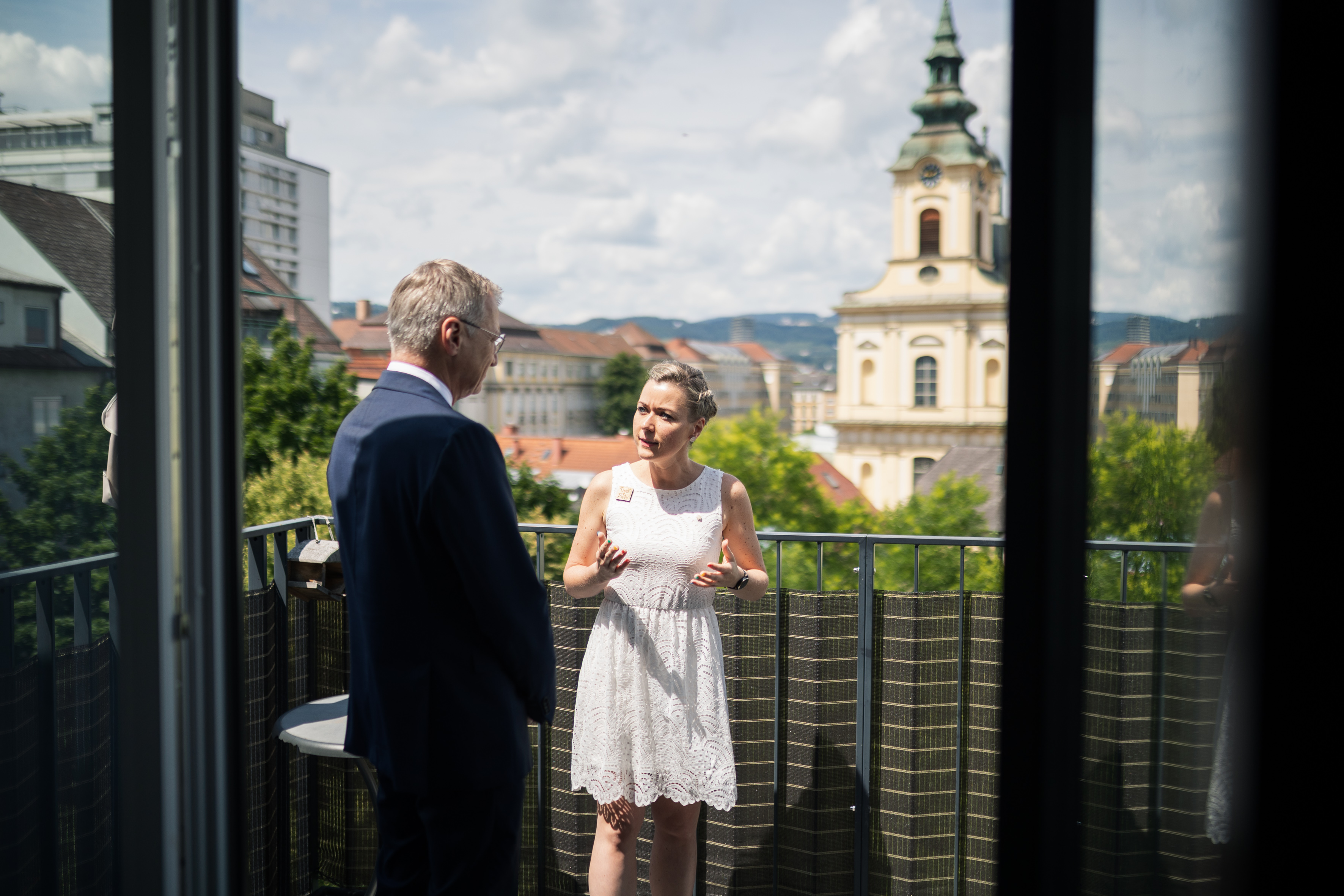 Das Bild zeigt den Landeshauptmann von hinten, während er mit einer blonden Frau in einem weißen Kleid spricht. Dabei stehen sie auf einem Balkon.